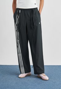 Fekete atlétikai nadrág fehér oldalsávokkal, húzózsinóros derékkal és laza fazonban. Világos rózsaszín cipőkkel párosítva. Sima textúra, hétköznapi dizájn.