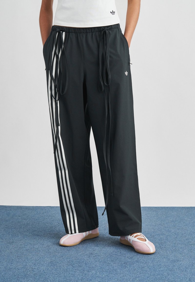 Fekete atlétikai nadrág fehér oldalsávokkal, húzózsinóros derékkal és laza fazonban. Világos rózsaszín cipőkkel párosítva. Sima textúra, hétköznapi dizájn.