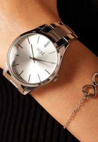 Silberne analoge Armbanduhr mit einem geb brushed Metallzifferblatt, dünnen Stundenmarkierungen und einem polierten Edelstahlarmband. Begleitet von einem silbernen Kettenarmband mit Herzanhänger.