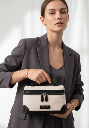 Woman in gray blazer holding a beige and black Diana & Co Firenze handbag with dual zipper and top handle against a light background.