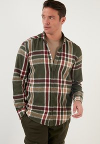 Camisa de franela verde con patrón a cuadros en rojo, blanco y marrón. Diseño de botones, mangas largas, textura suave y dobladillo redondeado.