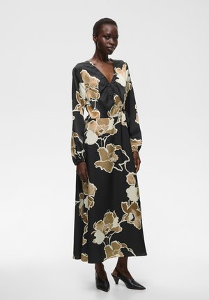 Femme portant une robe midi noire à manches longues avec un imprimé floral beige et blanc et des chaussures à talons noires à bout pointu.
