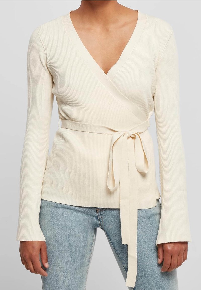 Femme portant un pull cache-cœur côtelé crème avec une ceinture à nouer, associé à un jean bleu clair, debout devant un fond uni.