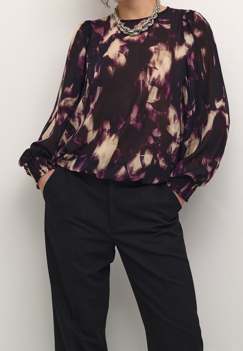 Blusa transparente con un patrón abstracto en púrpura y crema. Presenta mangas recogidas y puños elásticos, combinada con pantalones negros ajustados.