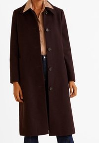 Dlouhý tmavě hnědý kabát z texturované látky, s předním zapínáním na knoflíky, bočními kapsami a špičatým límcem přes kostkovanou košili.