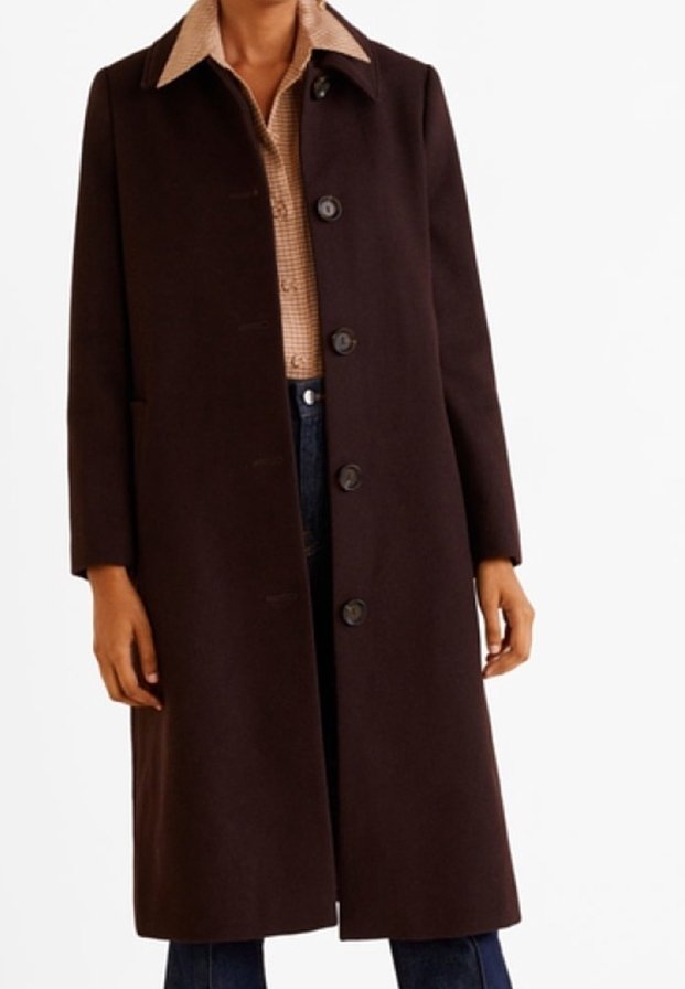 Dlouhý tmavě hnědý kabát z texturované látky, s předním zapínáním na knoflíky, bočními kapsami a špičatým límcem přes kostkovanou košili.