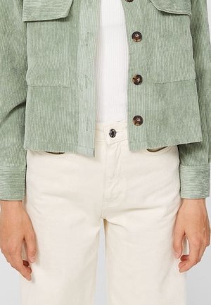 Veste en velours côtelé vert clair avec boutons marron portée sur un haut blanc côtelé et un pantalon crème taille haute.
