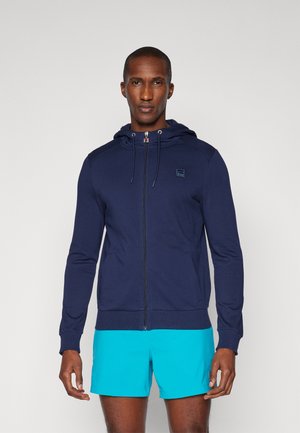 Homme portant un sweat à capuche zippé bleu marine et un short bleu vif, se tenant face à l'avant sur un fond uni.