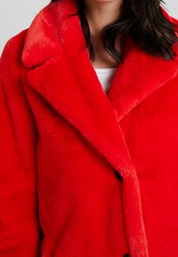 Abrigo de piel sintética roja con gran cuello de solapa con muesca, tejido texturizado y botones negros, mostrado en una persona.