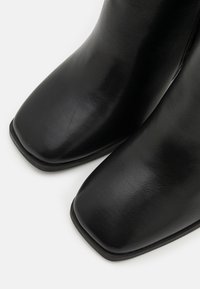 Bottes courtes en cuir noir avec une forme d'orteil carrée, une texture lisse et un design minimaliste. Fermeture éclair latérale pour faciliter l'enfilage.