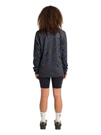 Osoba z kręconymi włosami, ubrana w czarną koszulę z długim rękawem z wzorem, czarne spodenki rowerowe z napisem na lewej nodze, białe skarpetki i czarne buty.