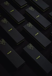 Sorte kasser med mat finish, prydet med et gult logo og designaccentueringer, arrangeret i et gitter på en mørk overflade.
