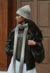 Black leather jacket with a brown fur collar, gray sweater, and a light gray patterned scarf. Gray and white knit beanie, holding a black bag.