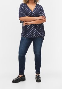 Kvinde står med armene over kors iført en navyblå bluse med prikker, mørkeblå skinny jeans og sorte platform loafers.