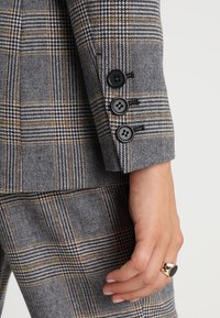 Manche de veste de costume à carreaux gris et bleu marine avec trois boutons noirs, une coupe ajustée, et un tissu texturé avec des accents dorés.