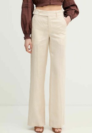 Frau trägt beige, hoch taillierte, weit geschnittene Hose, kombiniert mit einer bordeauxroten, kurz geschnittenen Bluse mit langen Ärmeln und braunen Sandalen mit offenen Zehen.