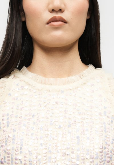 Primo piano della parte inferiore del viso e del collo di una donna che indossa una maglietta color crema decorata con paillettes iridescenti e scollo arricciato.