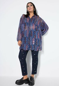 Mujer vestida con una túnica azul semitransparente de patrones, jeans oscuros con embellishments coloridos, zapatos de charol negros y aretes de aro frente a un fondo blanco.