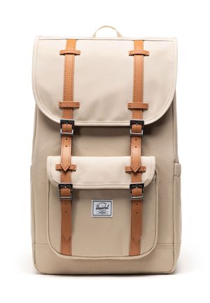 Mochila de lona beige con correas de cuero marrón y hebillas de metal, que cuenta con un bolsillo delantero con la etiqueta de la marca Herschel.