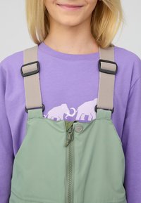 Grüne Latzhose mit zentralem Reißverschluss und verstellbaren beigen Trägern, getragen über einem lila Langarmshirt mit einem weißen Elefant-Motiv.