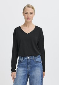 Schwarzes Langarmoberteil mit V-Ausschnitt, aus einem weichen Stoff gefertigt. Kombiniert mit hochgeschnittenen blauen Denim-Jeans mit sichtbaren Nähten.