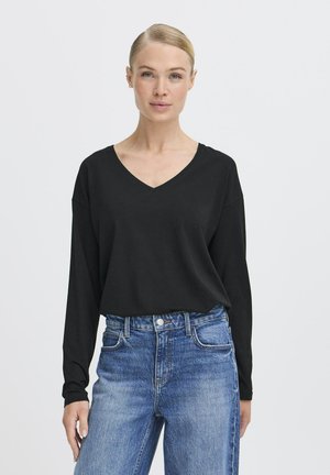 Schwarzes Langarmoberteil mit V-Ausschnitt, aus einem weichen Stoff gefertigt. Kombiniert mit hochgeschnittenen blauen Denim-Jeans mit sichtbaren Nähten.