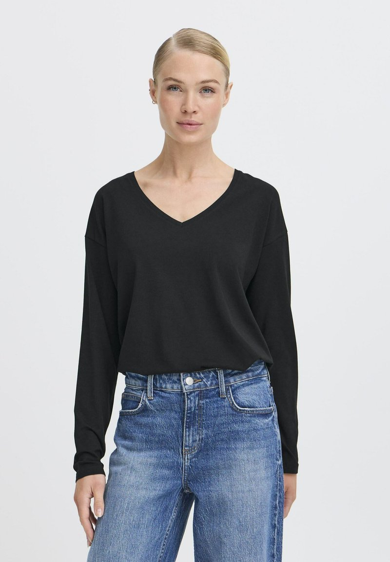 Schwarzes Langarmoberteil mit V-Ausschnitt, aus einem weichen Stoff gefertigt. Kombiniert mit hochgeschnittenen blauen Denim-Jeans mit sichtbaren Nähten.