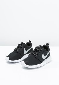 Une paire de chaussures de course Nike noires avec des semelles blanches et des logos swoosh gris, placées côte à côte sur une surface blanche.