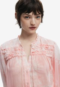 Blusa rosa con yoke arricciato, chiusura a bottoni frontale e una sottile texture a righe con motivo floreale. Materiale morbido e leggero.
