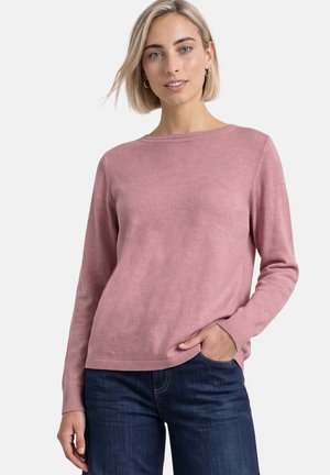 Femme aux cheveux blonds courts portant un pull rose à manches longues et un jean bleu foncé, posant avec une main dans la poche devant un fond blanc.