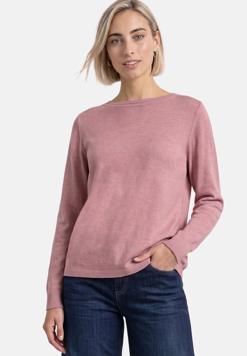 Femme aux cheveux blonds courts portant un pull rose à manches longues et un jean bleu foncé, posant avec une main dans la poche devant un fond blanc.