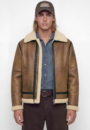 Nuori mies, joka käyttää ruskeaa shearling-takkia, farkkupaitaa, tummia housuja vyöllä ja ruskeaa lippalakkia, seisoo yksivärisen taustan edessä.