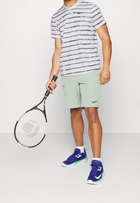 Grå och vit randig Nike-t-shirt med kort ärm, ljusgröna shorts med fickor, håller en svart och vit tennisklubba, blå och teal sneakers.