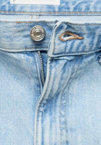 Jean en denim bleu clair avec une fermeture à bouton en métal et une fermeture éclair. Présente des coutures visibles et une passante de ceinture au niveau de la taille.