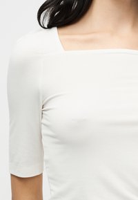 Λευκό, εφαρμοστό τοπ με τετράγωνη λαιμόκοψη και κοντά μανίκια. Κατασκευασμένο από μαλακό, ελαστικό ύφασμα με διακριτή υφή. Κατάλληλο για πολλές στιλιστικές επιλογές layering.