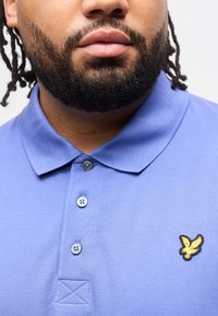 Ανδρας με μούσι και σκούρα μαλλιά που φοράει μπλε πόλο πουκάμισο με κουμπιά και έχει ένα κίτρινο λογότυπο αετού στο στήθος.