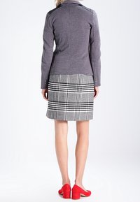 Chaqueta gris ajustada con cuello alto, combinada con una falda de cuadros en blanco y negro. Los zapatos de tacón bloque rojo completan el conjunto.