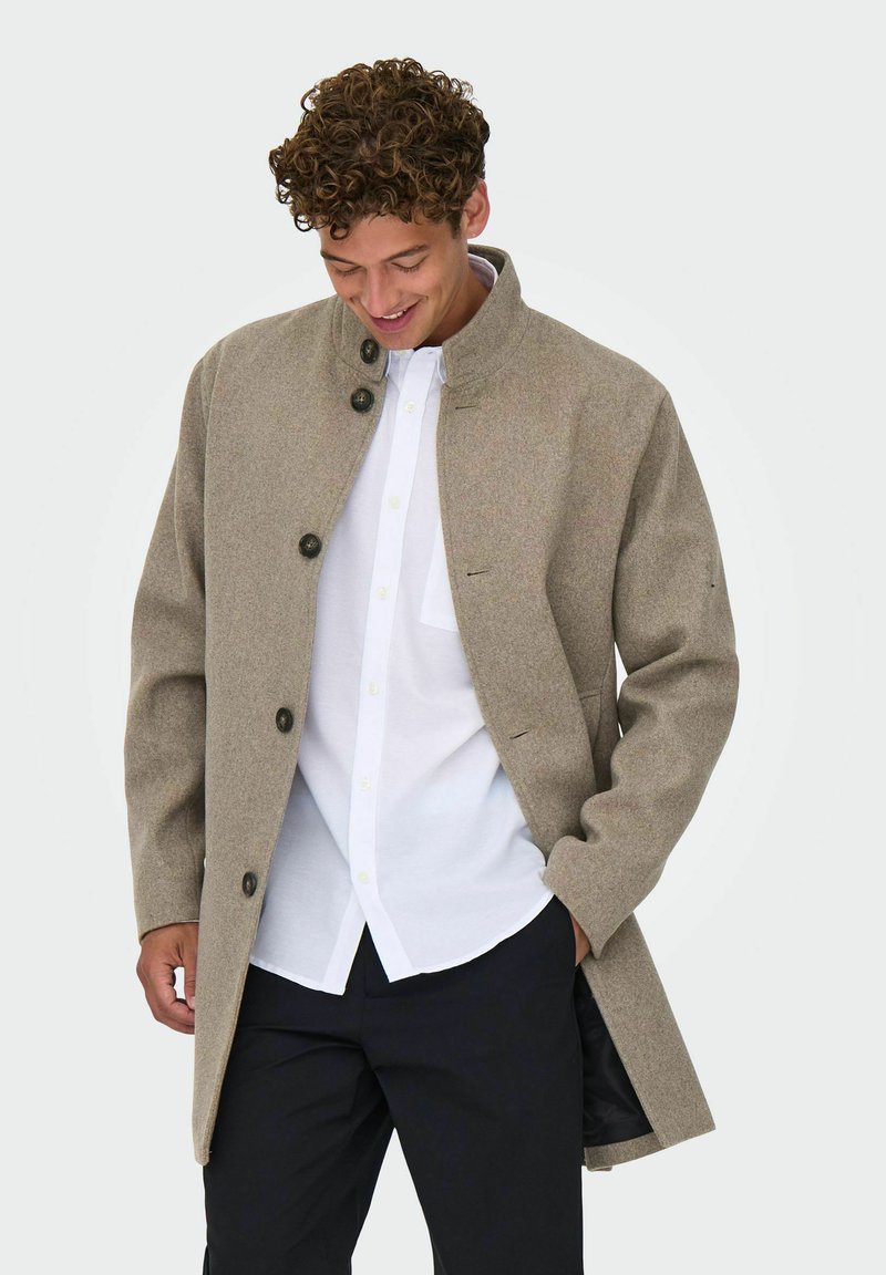 Brūns vilnas maisījuma mētelis ar augstu apkakli, pogu aizdari un garām piedurknēm. Nēsāts virs baltas krekla, ar elegantu, minimālistisku dizainu.