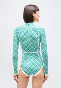 Γυναίκα με κοντά καστανά μαλλιά, φορώντας μαγιό μακ ρυμάνικο σε πράσινο και μπλε καρό με λευκή επένδυση, στέκεται μπροστά σε απλό φωτεινό φόντο.