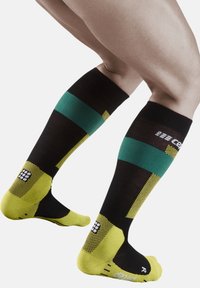 Calze nere fino al ginocchio con motivi verdi e gialli. Sezioni del piede e della caviglia testurizzate, con un design a scacchi e dettagli del logo.