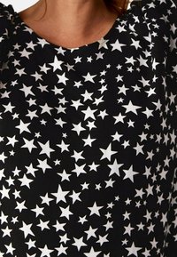 Czarny top z tkaniny z wzorem białych gwiazd w różnych rozmiarach, z zaokrąglonym dekoltem i marszczonymi detalami na ramionach.