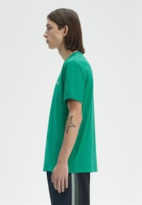 T-shirt in cotone verde con maniche corte, scollatura rotonda e dettaglio del logo sul davanti; mostrato su un modello di profilo, abbinato a pantaloni neri.