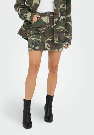 Femme portant une veste camouflage et une mini-jupe camouflage assortie avec des bottines noires, debout sur fond blanc.