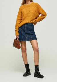 Grob gestrickter Senf-Pullover mit strukturierten Mustern. Dunkler Jeansrock mit Knöpfen vorne und Taschen. Schwarze Stiefeletten mit klobigen Absätzen.