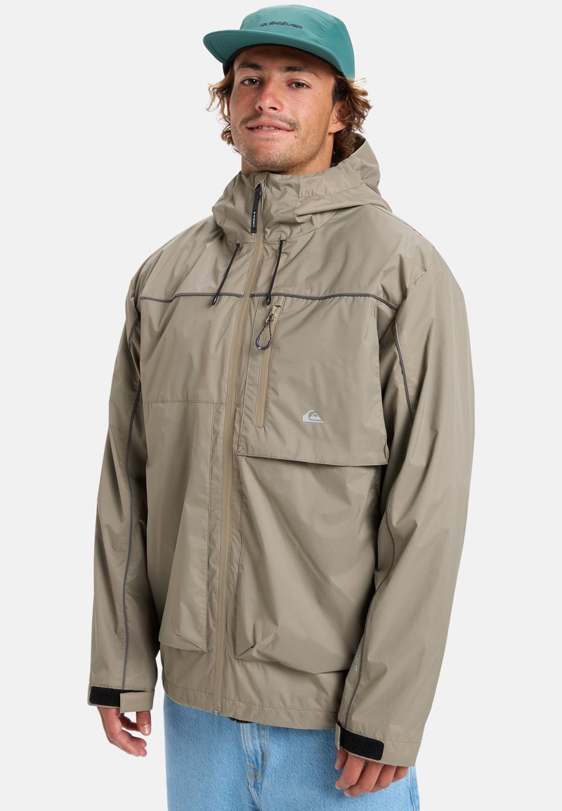 Beige waterdichte jack met ritssluiting aan de voorkant, verstelbare capuchon en zakken. Gekenmerkt door grijze stiksels en elastische manchetten met zwarte accenten.