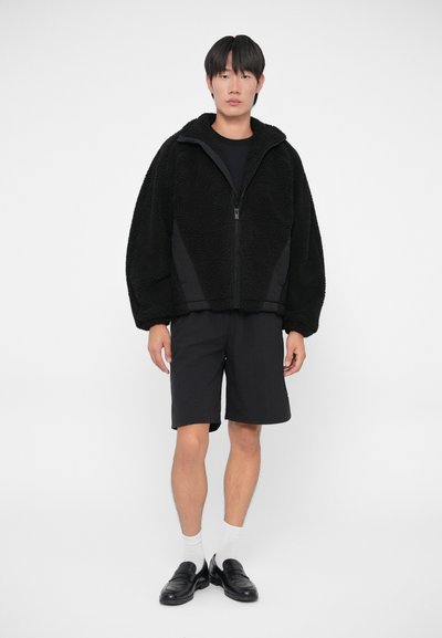 Zwarte fleecejack met grote kraag en rits, gecombineerd met zwarte shorts. Model draagt witte sokken en zwarte loafers. Glad, gestructureerd materiaal.