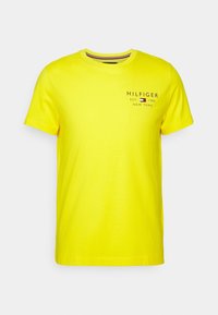 Žluté tričko s krátkým rukávem a kulatým výstřihem s malým černým a červeným logem a textem "Hilfiger Est 1985 New York" na levé horní části hrudi.