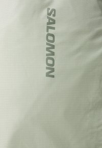 Světle zelená látka s jemnou texturou mřížky, s nápisem "SALOMON" v tmavě zelené barvě. Minimalistický design, bez dalších akcentů.