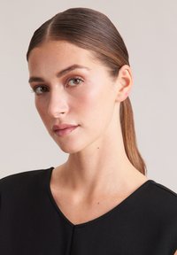 Vêtement noir avec un col en V, texture lisse et silhouette ajustée. Le modèle a les cheveux raides coiffés en arrière et un maquillage naturel.