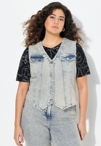 Chaleco de denim con un lavado azul claro, escote en forma de V y cierre de botones en la parte delantera. Presenta dos bolsillos en el pecho y un diseño ajustado, combinado con una camiseta negra de patrón.